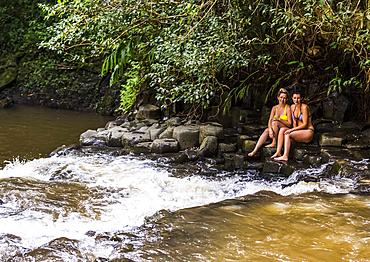 Two women wearing bikinis sitting at one of the Twin Falls waterfalls and looking at the camera; Haiku, Maui, Hawaii, United States of America