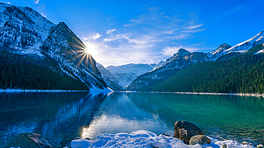 Lake Louise reflecting the Rocky Mountains with a sunburst shining behind a mountain and snow in autumn, Banff National Park; Alberta, Canada