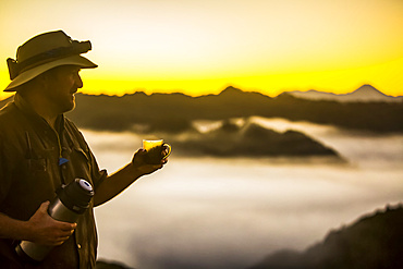 The Blue Duck lodge located in the Whanganui National Park is a working cattle farm with a focus on conservation. Travellers go to a scenic viewpoint to watch the sunrise over the rainforest. A man stands drinking coffee and holding a thermos; Retaruke, Manawatu-Wanganui, New Zealand