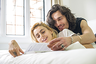 A cool young couple lying on a bed looking at a book.