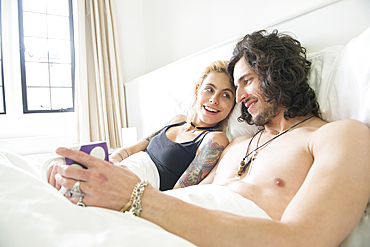 A cool young tattooed couple lying in a bed enjoying a cup of tea.