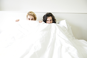 A cool young tattooed couple lying in a double bed playing peekaboo with the camera.