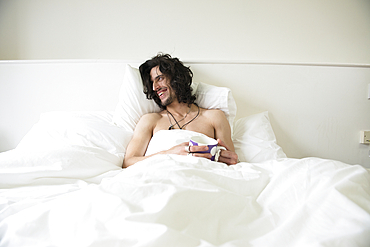 A young man with long hair lying in a bed with a cup of tea looking happy.