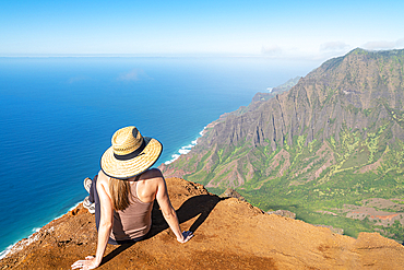 View taken from behind of a woman in a straw hat sitting on the top of a mountain cliff looking out onto the brilliant, blue water of the Pacific Ocean and mountainous coastline along the Kalalau Trail on the Napali Coast, Kauai, Hawaii, United States of America