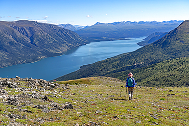 View taken from behind of a person walking down a mountainside with a spectacular view of the Kusawa Lake below, Yukon, Canada