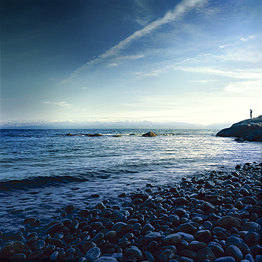 Person stands on a rocky slope looking out to the water along the tranquil coast, Alaska, United States of America