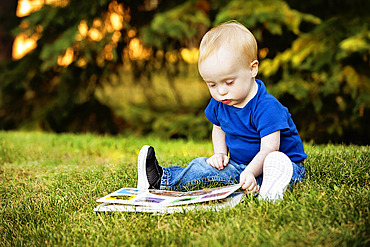 Baby boy with Down syndrome sitting and looking at a picture book on grass in a city park during a warm fall afternoon, Leduc, Alberta, Canada