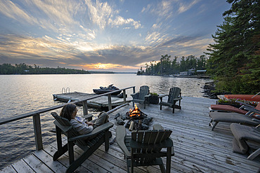 Woman sits in an adirondack chair on a lakeside deck in front of a fire pit, watching the sunset in the distance, Lake of the Woods, Ontario, Canada