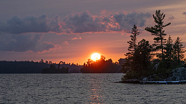 Dramatic view of the warm glowing sun sinking over the horizon with a lake and silhouetted trees along the shoreline, Lake of the Woods, Ontario, Canada