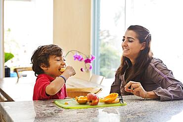 Hispanic mother and son eating fruit in kitchen