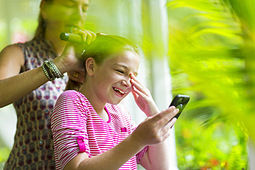 Caucasian mother brushing hair of daughter texting on cell phone