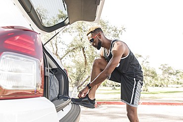 Black man leaning on car hatchback tying shoe