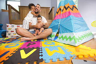 Father sitting on playroom floor holding son and daughter