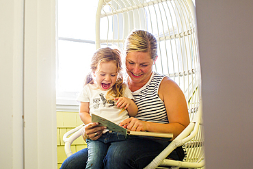 Caucasian mother reading book to excited daughter sitting on lap