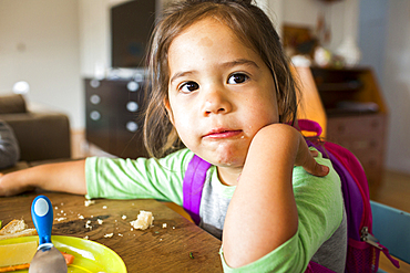 Messy mixed race girl eating food and wearing backpack at table