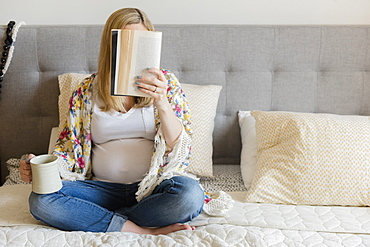 Caucasian expectant mother sitting on bed reading book and drinking coffee