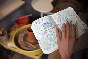 Hand of Caucasian woman examining drawings in book near pottery wheel