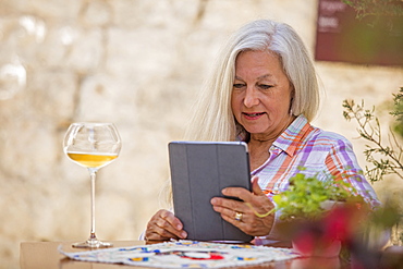 Older Caucasian woman drinking white wine and using digital tablet