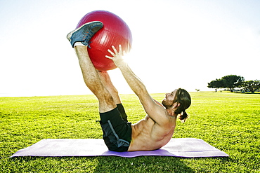 Caucasian man lifting fitness ball with legs in sunny field