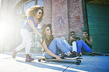 Woman pushing friend sitting on skateboard in city