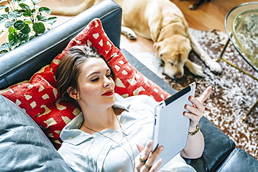 Woman laying on sofa near dog using digital tablet