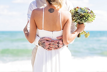 Caucasian bride and groom with tattoos hugging on beach