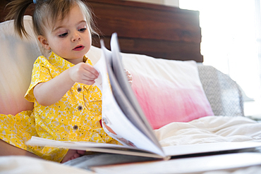 Caucasian baby girl sitting on bed reading book