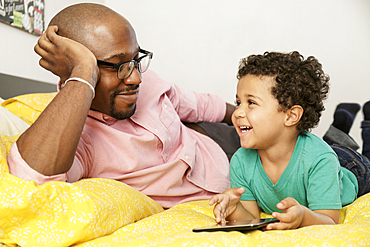 Father watching son laying on bed using digital tablet