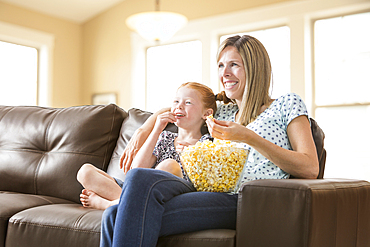 Caucasian mother and daughter sitting on sofa eating bowl of popcorn