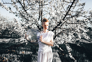 Unusual Caucasian teenage girl holding glass helmet near tree