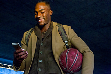 Black man holding basketball texting on cell phone