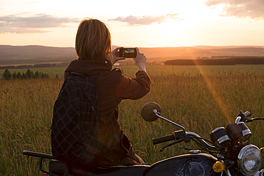 Caucasian woman with motorcycle in field photographing sunset