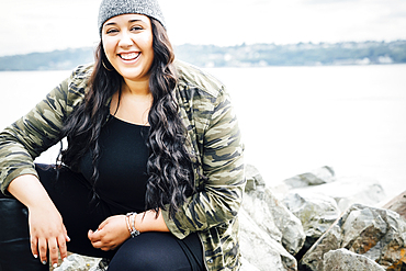 Smiling Mixed Race woman sitting on rocks near ocean