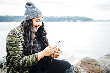 Smiling Mixed Race woman texting on cell phone near ocean