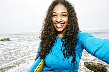 Smiling Mixed Race woman holding surfboard posing for selfie at beach