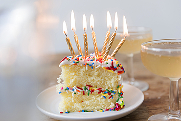 Candles burning on slice of cake with sprinkles near champagne