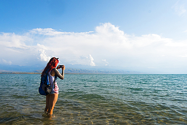 Caucasian woman wading in ocean photographing with cell phone