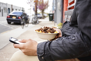 Hispanic man eating bowl of food on sidewalk using cell phone