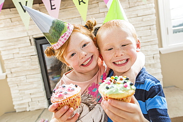 Caucasian boy and girl wearing party hats showing cupcakes