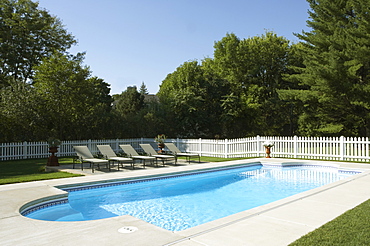 Overall of pool and surrounding area, white picket fence, 5 chaise lounges in a row by pool. Rectangular shape w/half circles at each end. Trees in background