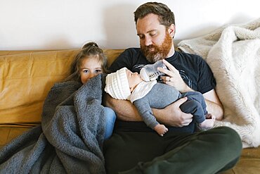 Girl (2-3) assisting father feeding newborn son (0-1 months) on sofa