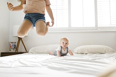 Girl (2-3) jumping on bed with baby brother (6-11 months) in background