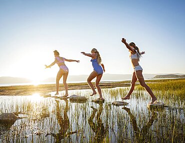 Girls (10-11, 12-13, 14-15) walking on stepping stones in lake at sunrise