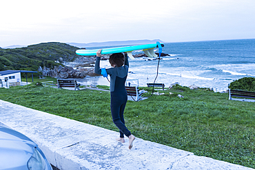 South Africa, Hermanus, Boy (10-11) carrying surfboard on head while walking on sidewalk at dusk in Voelklip Beach