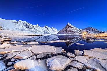 Star trails and lights on the snowy peaks reflected in the cold sea, Volanstinden, Fredvang, Lofoten Islands, Northern Norway, Scandinavia, Europe