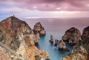 Photographer on top of cliffs surrounded by sea under the pink sky at sunrise, Ponta Da Piedade, Lagos, Algarve, Portugal, Europe