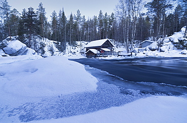 Dusk frames the frozen water in the snowy woods and a wooden hut, Juuma, Myllykoski, Lapland region, Finland, Europe