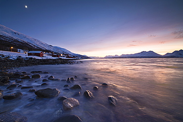 Fiery sky at sunset on typical Rorbu framed by snowy peaks and frozen sea, Djupvik, Lyngen Alps, Troms, Norway, Scandinavia, Europe