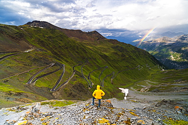 Man on rocks admires the rainbow on the winding road, Stelvio Pass, South Tyrol side, Valtellina, Lombardy, Italy, Europe