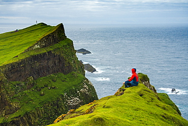 Man sitting on cliffs looks towards the lighthouse, Mykines island, Faroe Islands, Denmark, Europe